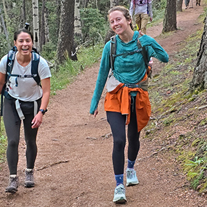 Two women hiking on a trail in the woods with backpacks on